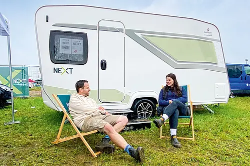 Ein Paar entspannt in Liegestühlen vor einem weißen NEXT Caravan auf einer grünen Wiese beim Camping Urlaub im Freien