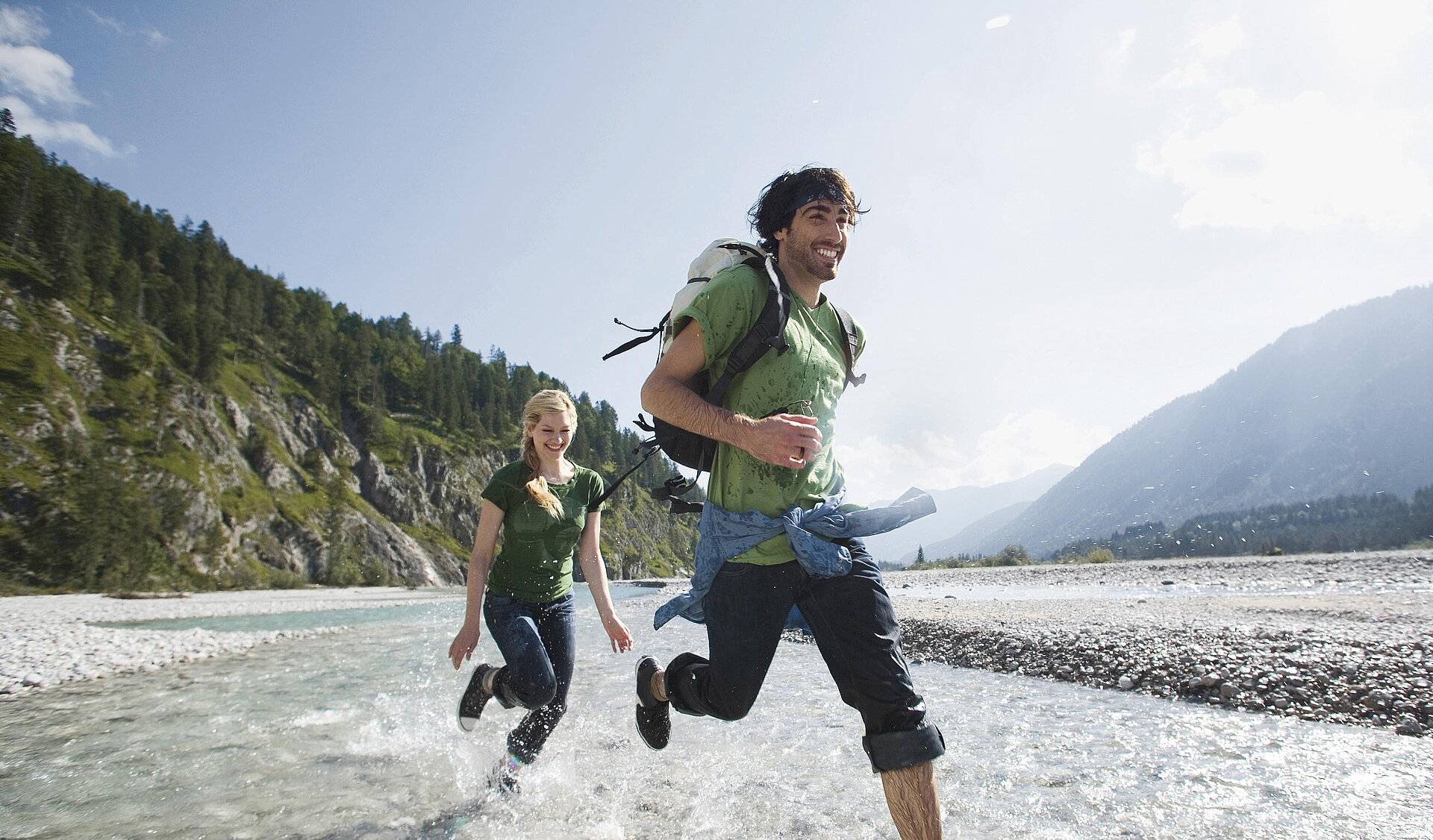 Junges Paar rennt lachend durch einen seichten Gebirgsfluss beim Aktivurlaub in den Bergen mit dem NEXT Caravan