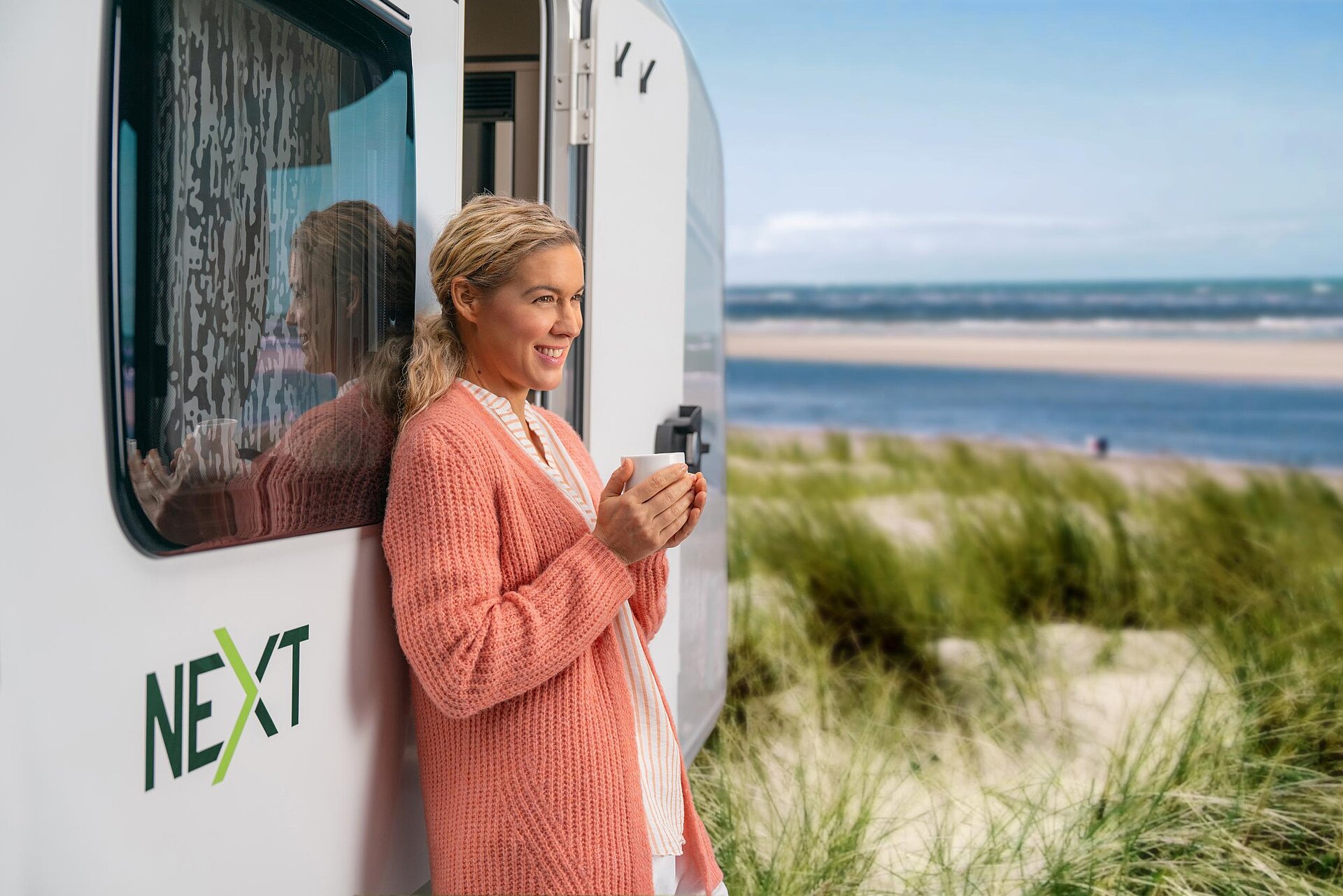 Seitenansicht eines NEXT Caravans mit Logo und einer Frau die bei einer Tasse Kaffee die Aussicht auf den Ozean genießt