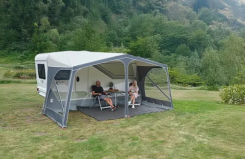 Ein Paar sitzt entspannt an einem Tisch unter dem grauen Vorzelt eines NEXT Caravans auf einer grünen Wiese im Wald