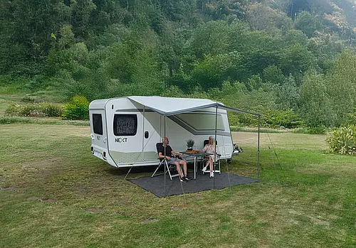Ein Paar sitzt entspannt an einem Tisch unter der grauen Markise ihres NEXT Caravans auf einer grünen Wiese im Wald