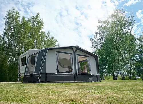 Weißer NEXT Caravan mit großem grau-weißen Vorzelt und Gardinen auf einer grünen Wiese vor Birken bei Sonnenschein