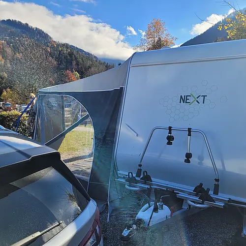 NEXT Caravan mit angebautem Vorzelt und Fahrradträger auf einem Campingplatz vor bewaldeten Bergen und blauem Himmel