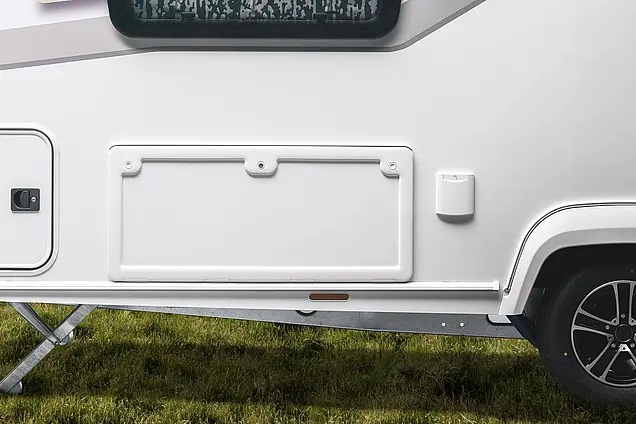 Seitenansicht der geschlossenen weißen Serviceklappe am Heck des NEXT Caravan über dem Fahrgestell auf einer Rasenfläche