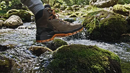 Wanderer mit braunem Bergschuh überquert einen klaren Bachlauf über moosige Steine beim Outdoor Urlaub mit dem NEXT Caravan